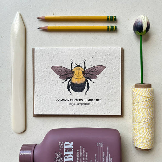 Card with a bee illustration on a light surface with stationery items.