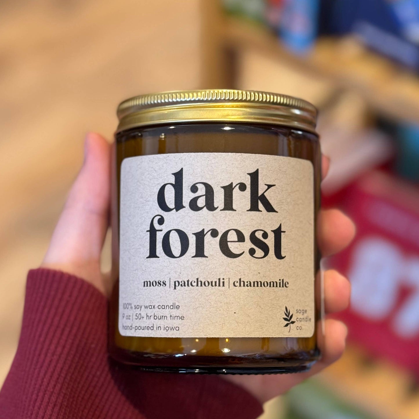 A hand wearing a dark red sweater holds a glass jar candle labeled 'dark forest' in a softly blurred indoor setting with bookshelves in the background.