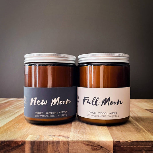 Two candles labeled 'New Moon' and 'Full Moon' on a wooden surface with a dark background.