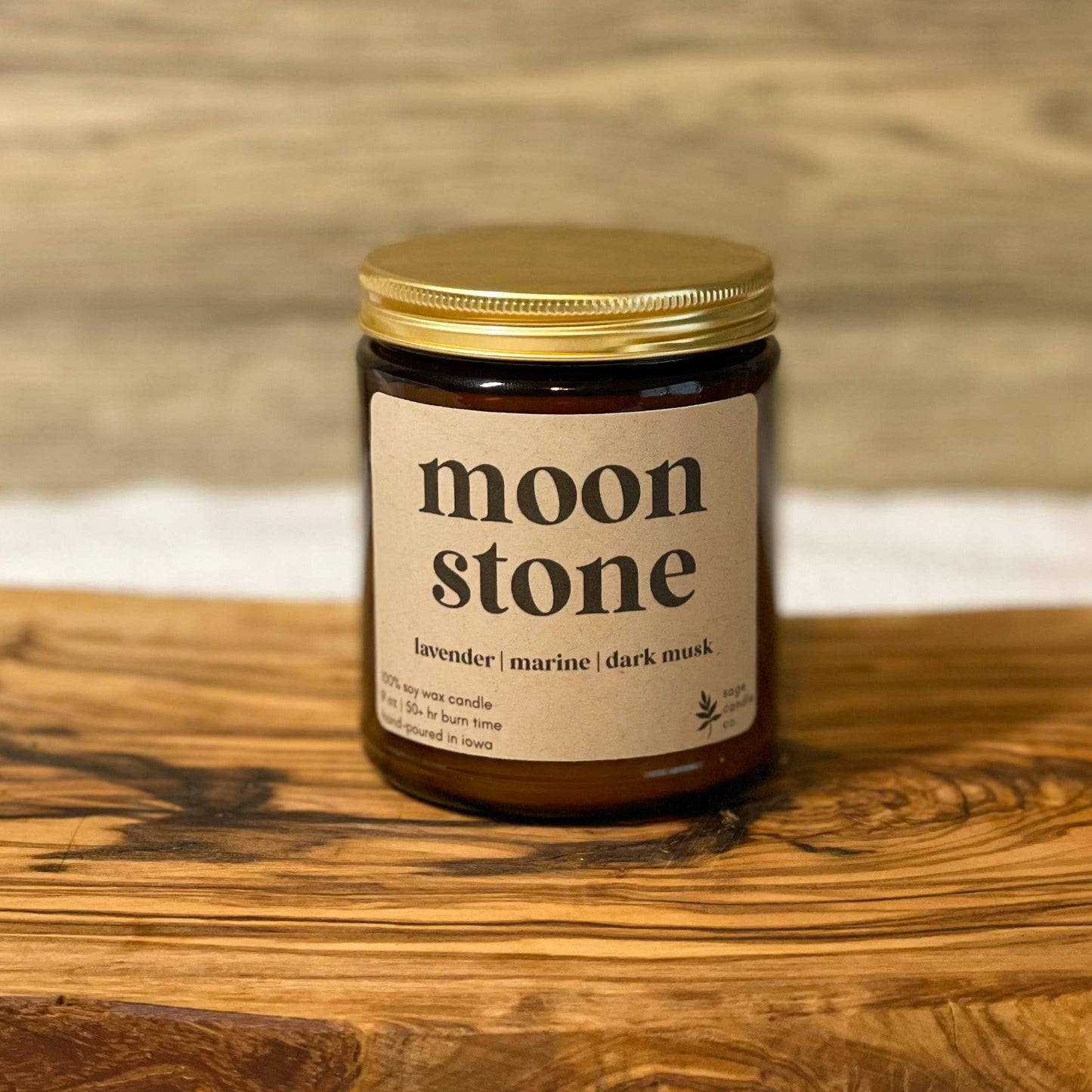 A jar candle labeled 'moon stone' with a gold lid and cream label, placed on a rustic wooden board against a light wood wall background.