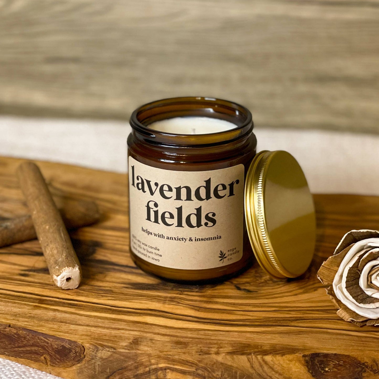 An amber glass jar candle labeled 'lavender fields' sits open on a rustic wooden board, with its gold lid beside it and a rolled paper flower and cinnamon stick as styled props against a wood plank background.