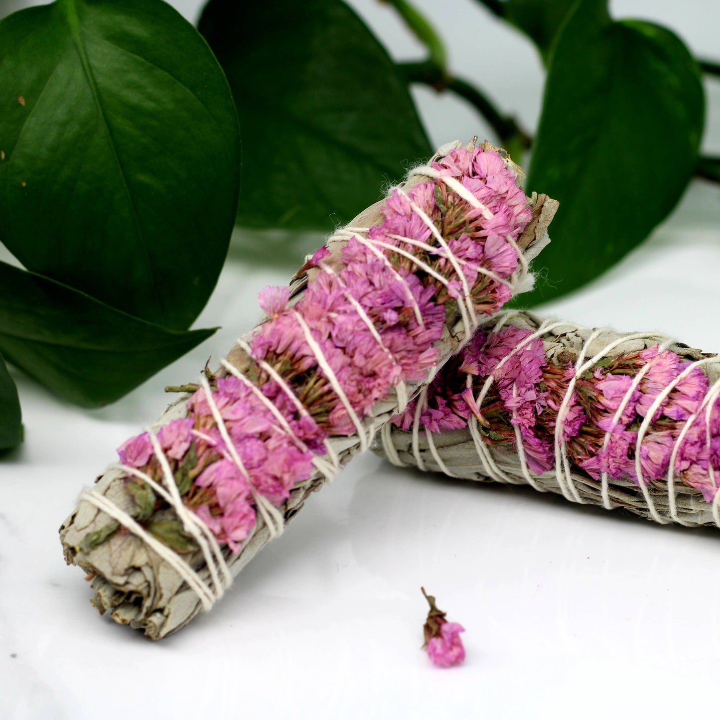 Two rolled sage bundles with pink flowers and white string on a white surface with green leaves in the background.