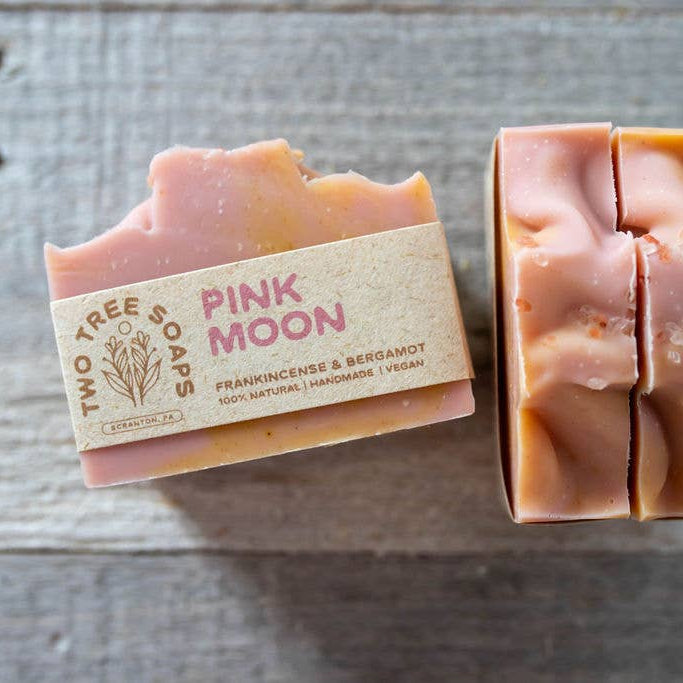 Two bars of soap, one with packaging labeled 'Pink Moon' and 'Two Tree Studio', on a wooden surface.