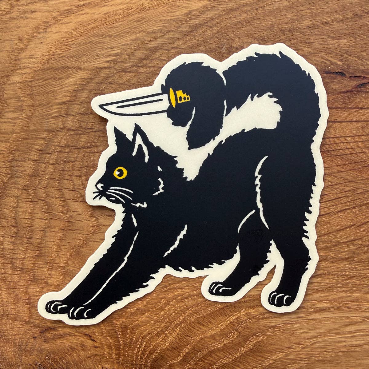 A die-cut vinyl sticker featuring a black cat with a knife in its mouth, shown on a warm brown wood grain background.