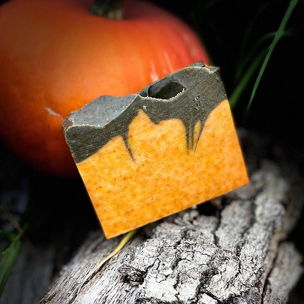 A rectangular orange and brown decorative plaque or tile with a cutout design, propped against a textured log with a small orange pumpkin in the background against a black backdrop.