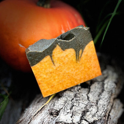 A rectangular orange and brown decorative plaque or tile with a cutout design, propped against a textured log with a small orange pumpkin in the background against a black backdrop.