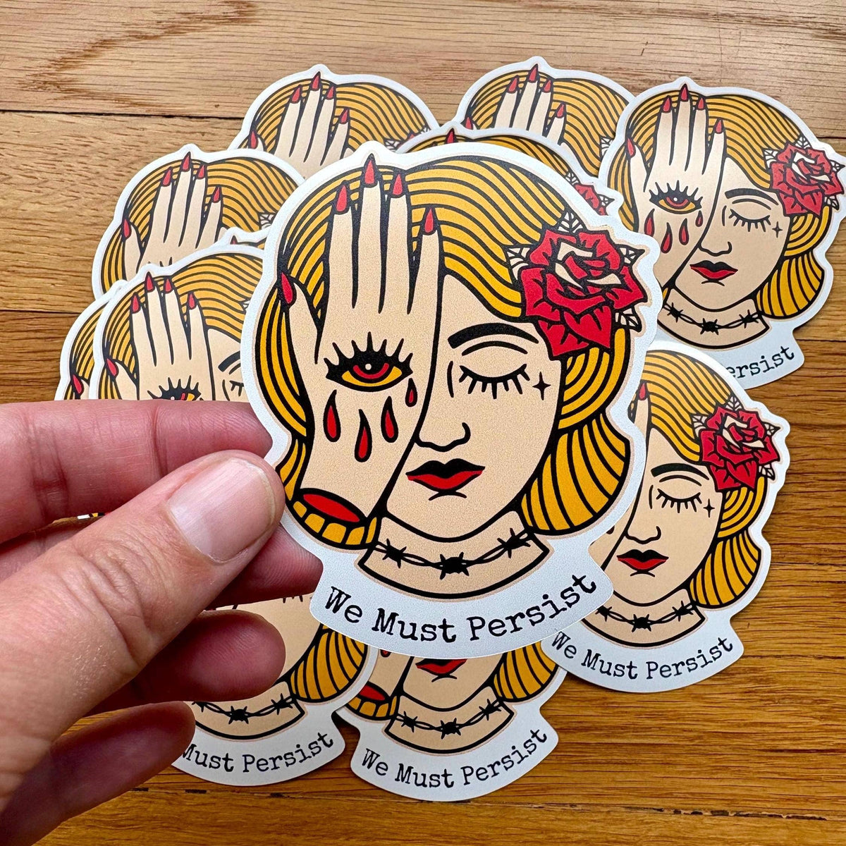 A hand holds a die-cut sticker in front of a stack of identical stickers on a wooden surface. The sticker design shows a stylized blonde woman with closed eyes, a hand over one face, red tears, a red rose, and the text 'We Must Persist'.