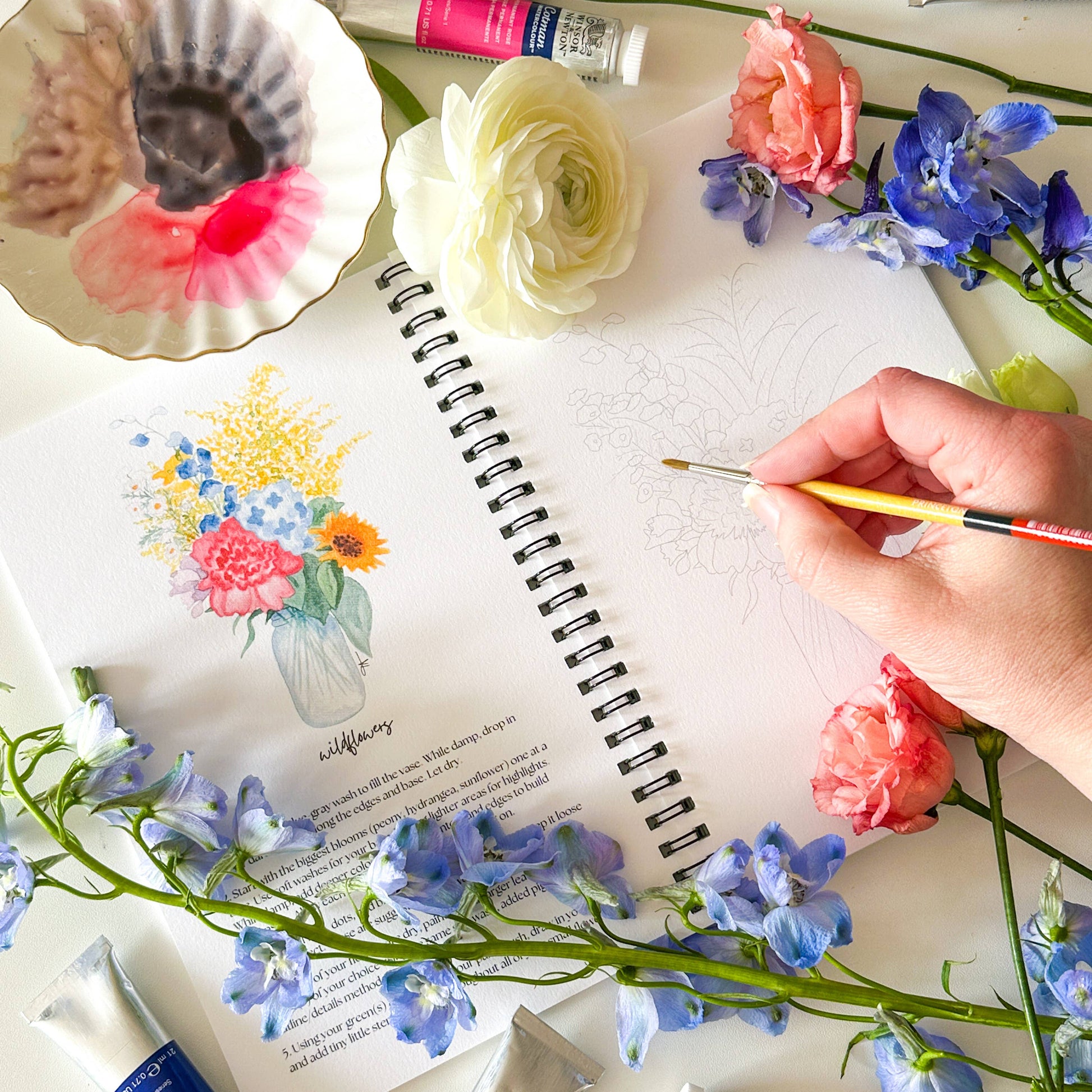 A hand holding a paintbrush sketches in a spiral-bound art notebook, surrounded by watercolor paints, flowers, and a palette on a white surface.
