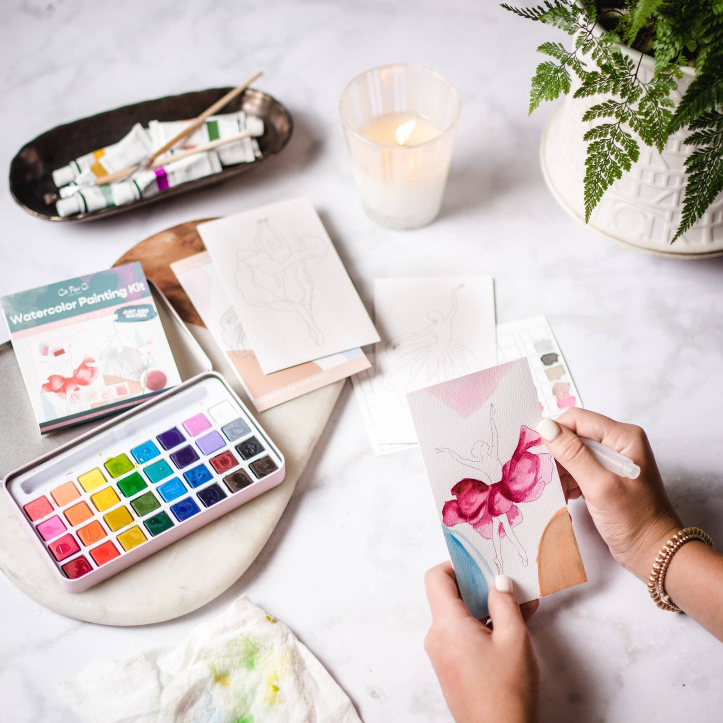 Person holding a watercolor painting with a set of watercolors and art supplies on a table.