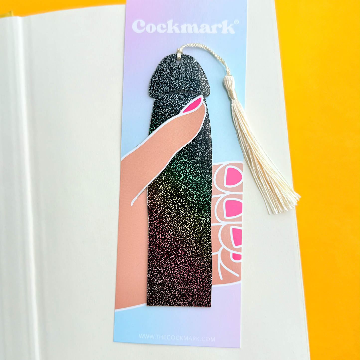 A novelty bookmark shaped like a glittery dildo with a cream tassel, shown resting on a white book page against a bright yellow background. The bookmark features a black-to-green glitter gradient body, pink nail graphics, and the brand name 'Cockmark' printed at the top.