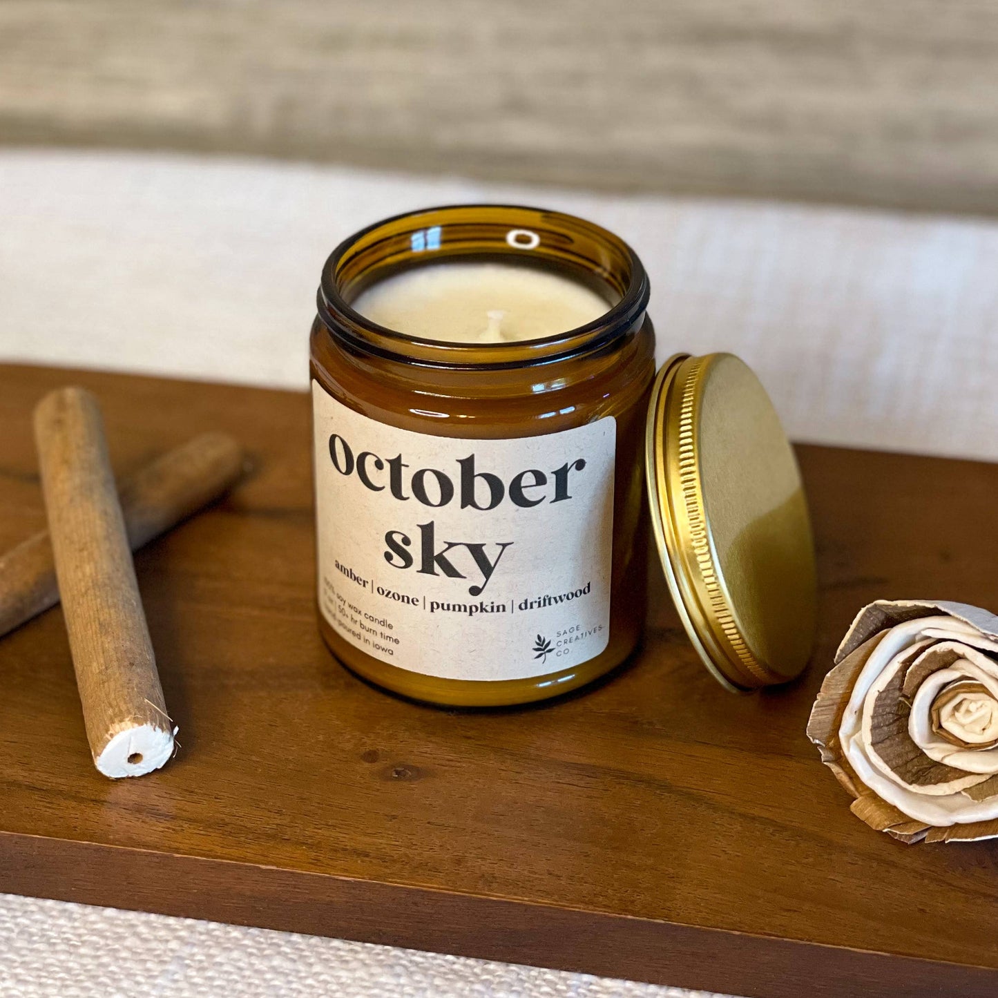 An amber glass jar candle labeled 'october sky' with its gold lid off, placed on a wooden board with two rolled papers and a paper flower against a light wood background.