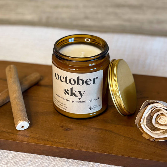 An amber glass jar candle labeled 'october sky' with its gold lid off, placed on a wooden board with two rolled papers and a paper flower against a light wood background.
