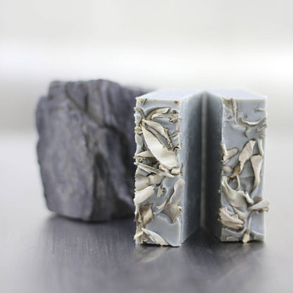 A handmade soap bar with a light gray base and embedded dried botanical pieces, shown in front of a dark gray stone on a reflective surface.