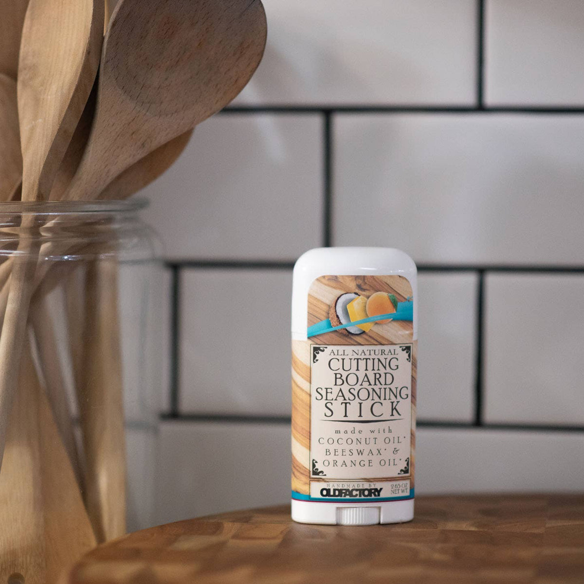 A stick of Cleary All Natural Cutting Board Seasoning Stick standing on a wooden surface, with a blurred kitchen background featuring white subway tiles and wooden utensils in a glass jar.