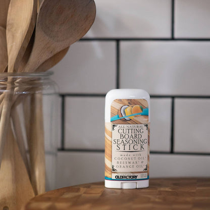 A stick of Cleary All Natural Cutting Board Seasoning Stick standing on a wooden surface, with a blurred kitchen background featuring white subway tiles and wooden utensils in a glass jar.