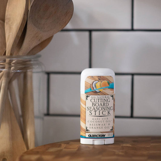 A stick of Cleary All Natural Cutting Board Seasoning Stick standing on a wooden surface, with a blurred kitchen background featuring white subway tiles and wooden utensils in a glass jar.