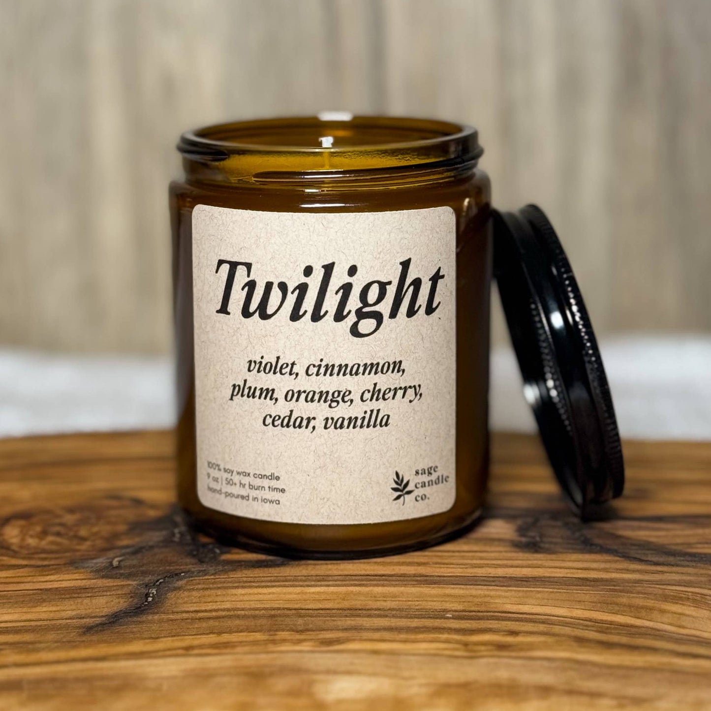 An amber glass jar candle labeled 'Twilight' with its black lid resting beside it, set on a rustic wood slab with a blurred wood-paneled background.