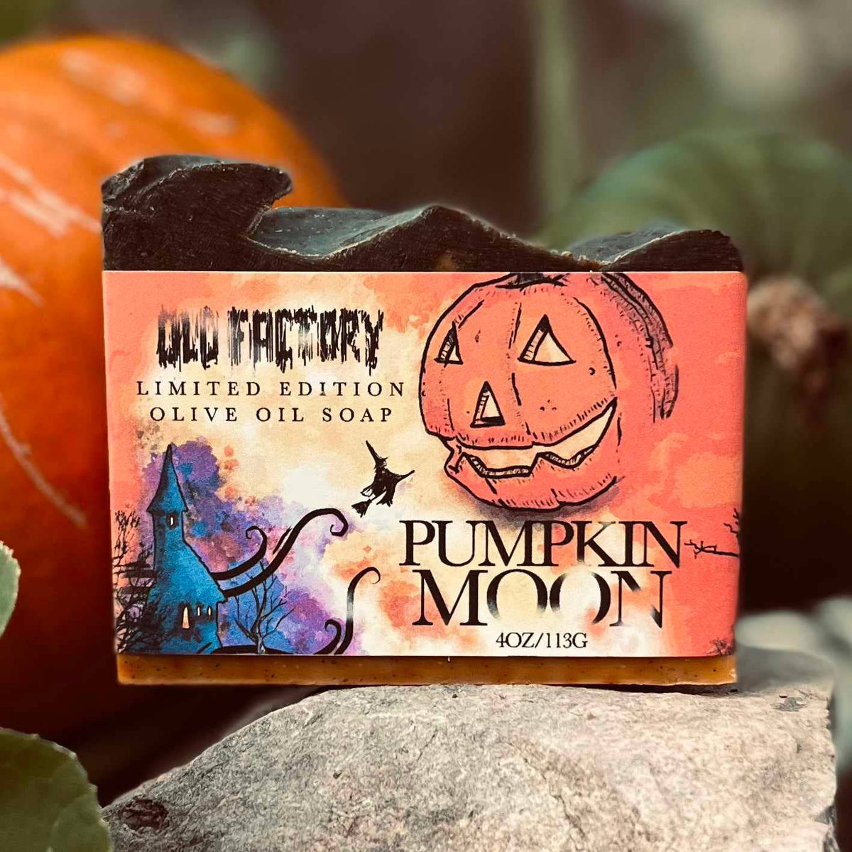 A bar of Old Factory Limited Edition 'Pumpkin Moon' olive oil soap, styled on a stone with pumpkins in the background. The soap has a dark, textured top and a colorful orange, red, and purple label featuring a jack-o'-lantern and a haunted house.