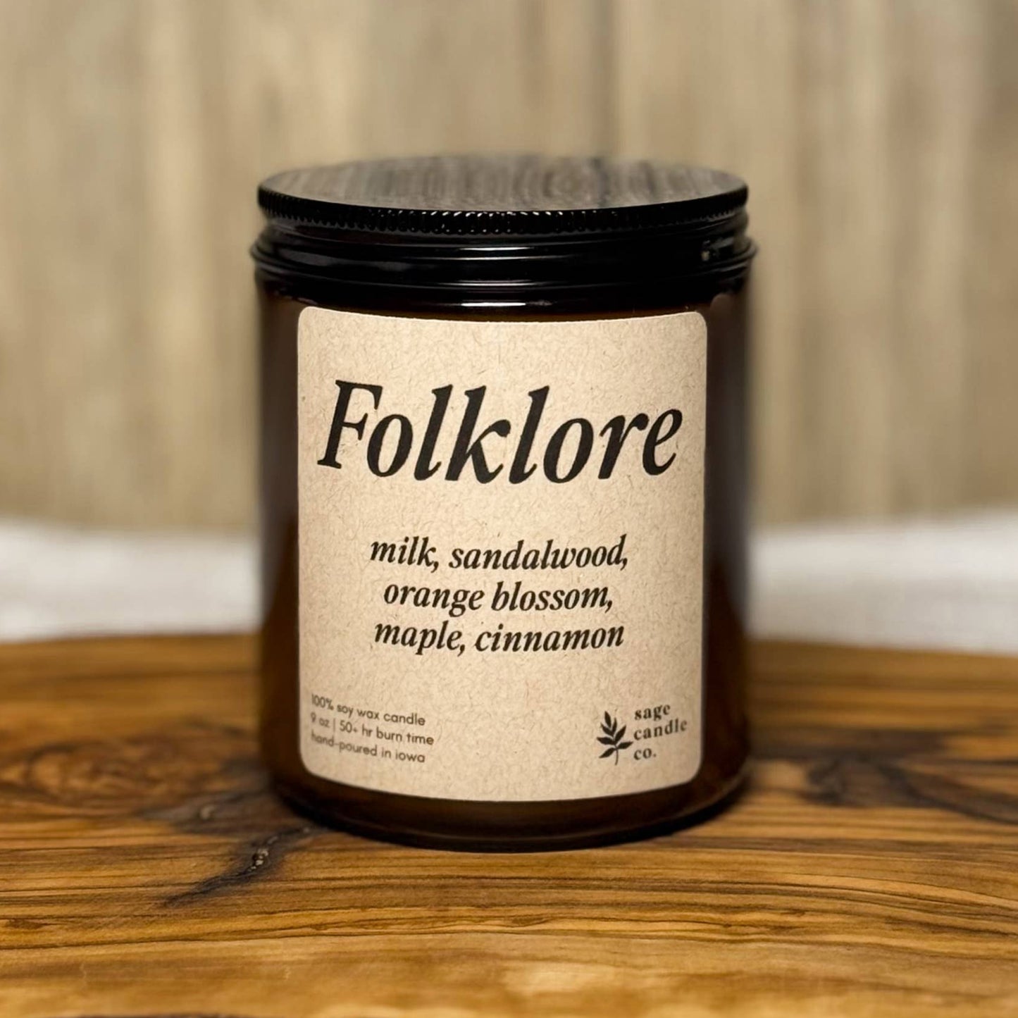 A dark amber glass candle jar with a black lid and a cream label reading 'Folklore' and 'milk, sandalwood, orange blossom, maple, cinnamon,' placed on a rustic wood slab with a light wood wall background.