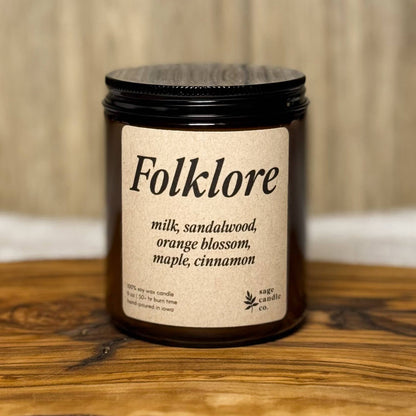 A dark amber glass candle jar with a black lid and a cream label reading 'Folklore' and 'milk, sandalwood, orange blossom, maple, cinnamon,' placed on a rustic wood slab with a light wood wall background.