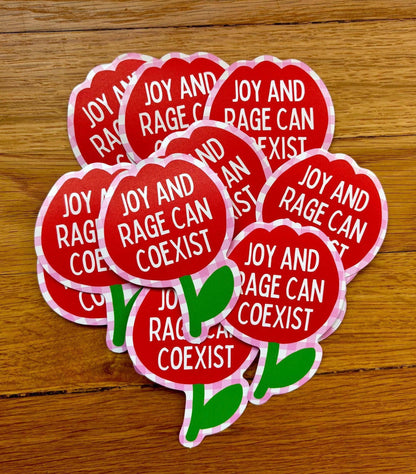Red stickers with 'Joy and Rage can coexist' text on a wooden surface