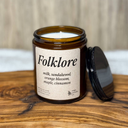 An amber glass jar candle labeled 'Folklore' with its black lid resting beside it on a rustic wood slab, set against a light wood paneled background.