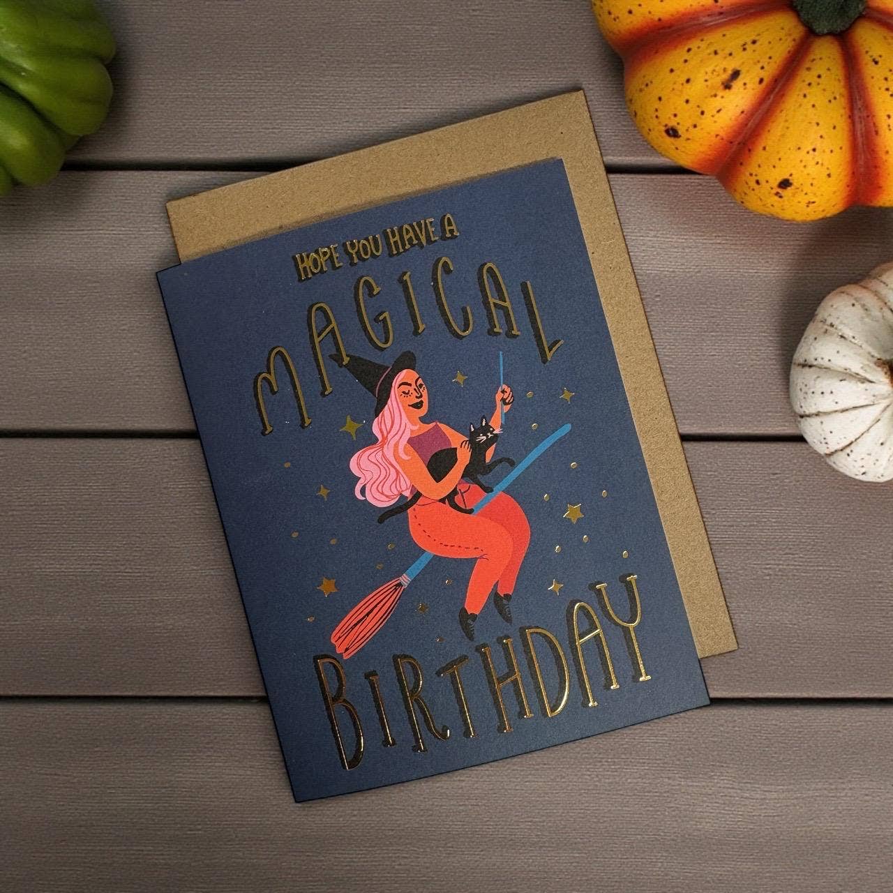 Birthday card with witch illustration and text on a wooden surface with pumpkins.