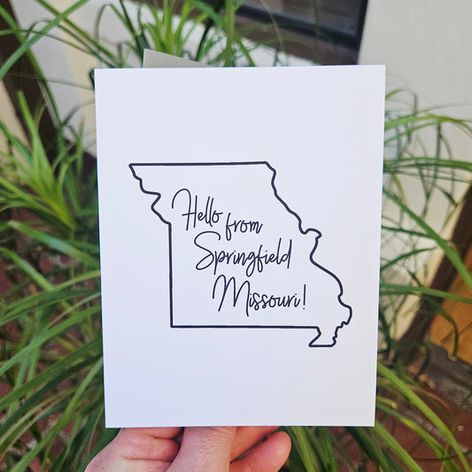 Card with outline of Missouri and 'Hello from Springfield, Missouri!' text held by a hand with plants in the background