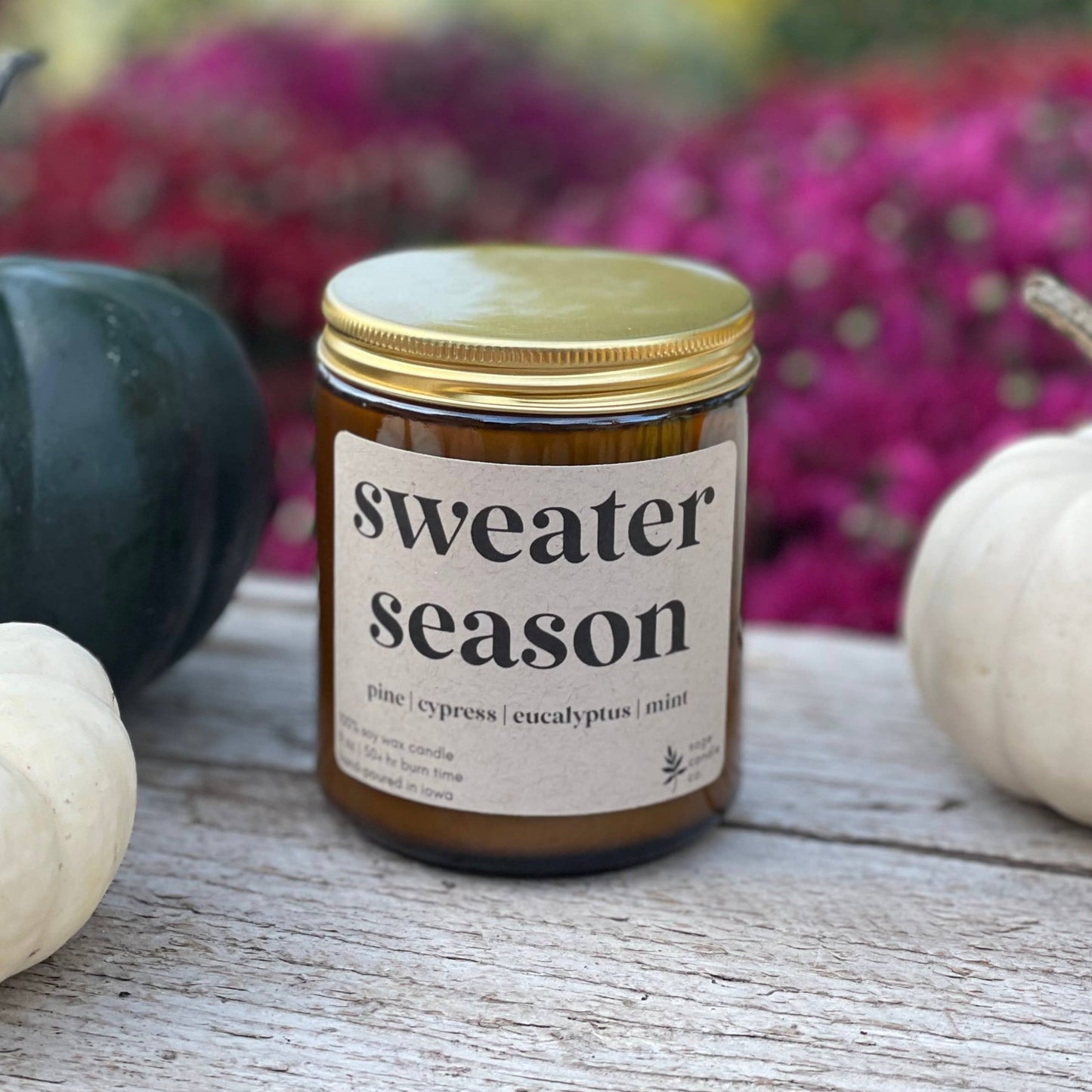 A candle in an amber glass jar with a gold lid and a cream label reading 'sweater season' sits on a rustic wooden surface, flanked by white and dark green pumpkins with a blurred background of colorful flowers.