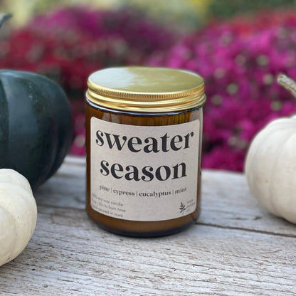 A candle in an amber glass jar with a gold lid and a cream label reading 'sweater season' sits on a rustic wooden surface, flanked by white and dark green pumpkins with a blurred background of colorful flowers.