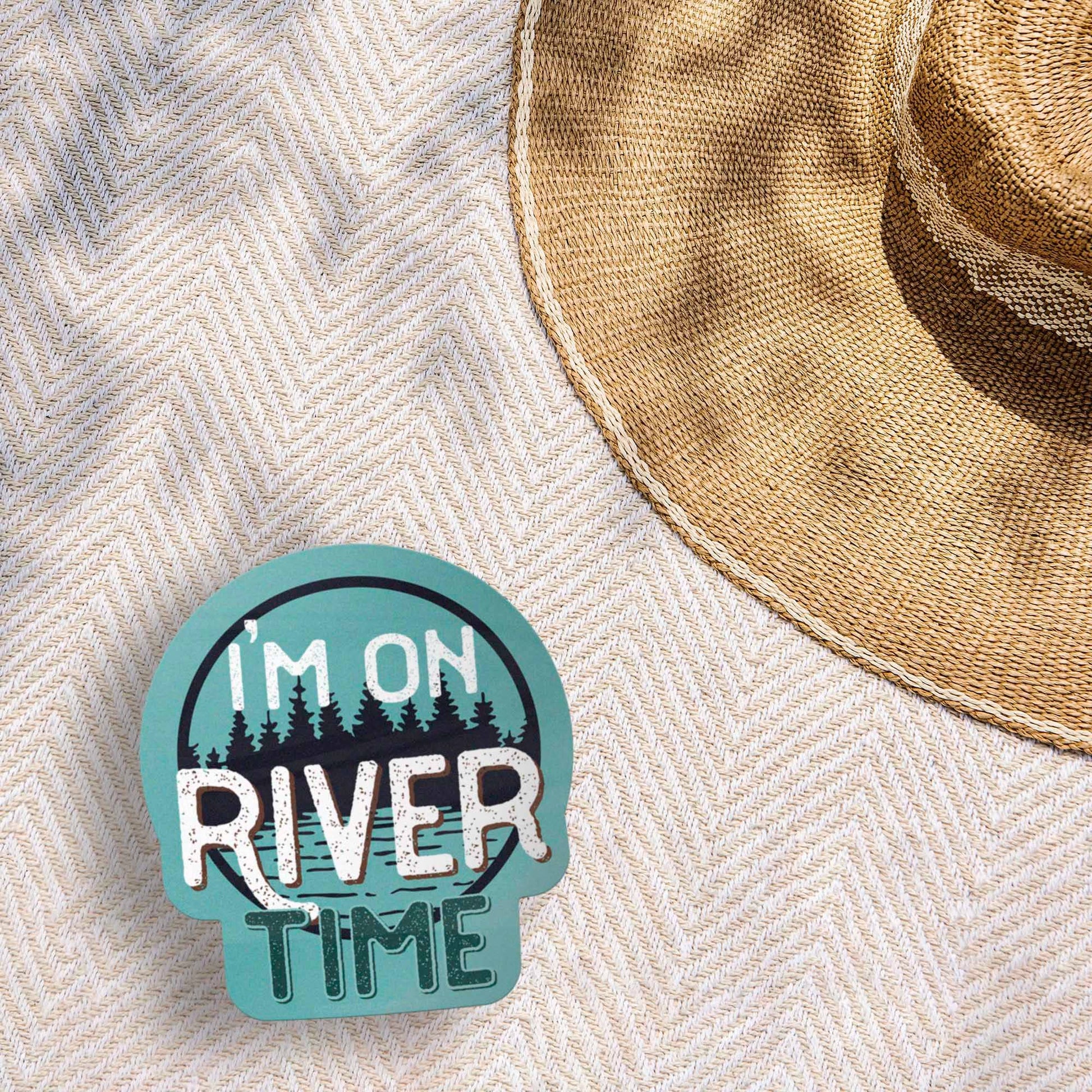 Sticker with 'I'm on River Time' text on a textured surface with a straw hat.