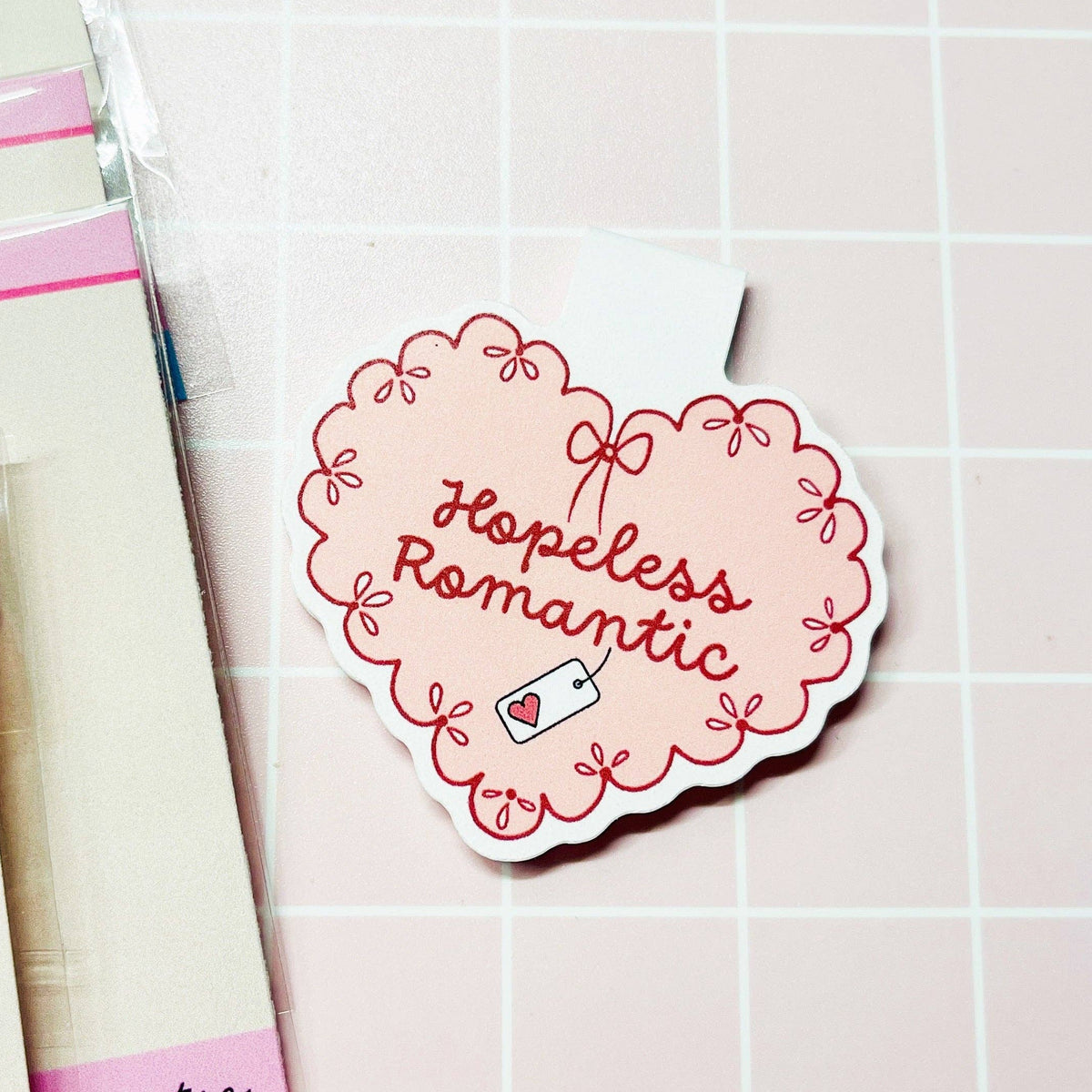 A heart-shaped sticker with the phrase 'Hopeless Romantic' in red script, surrounded by a pink and red scalloped border, displayed on a light pink grid background next to packaged stickers.