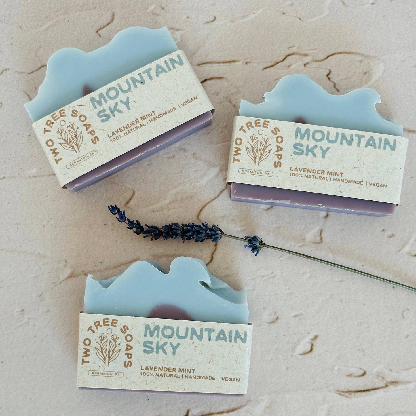 Three bars of 'Mountain Sky' soap from Two Tree Soaps on a textured beige surface with lavender flowers.