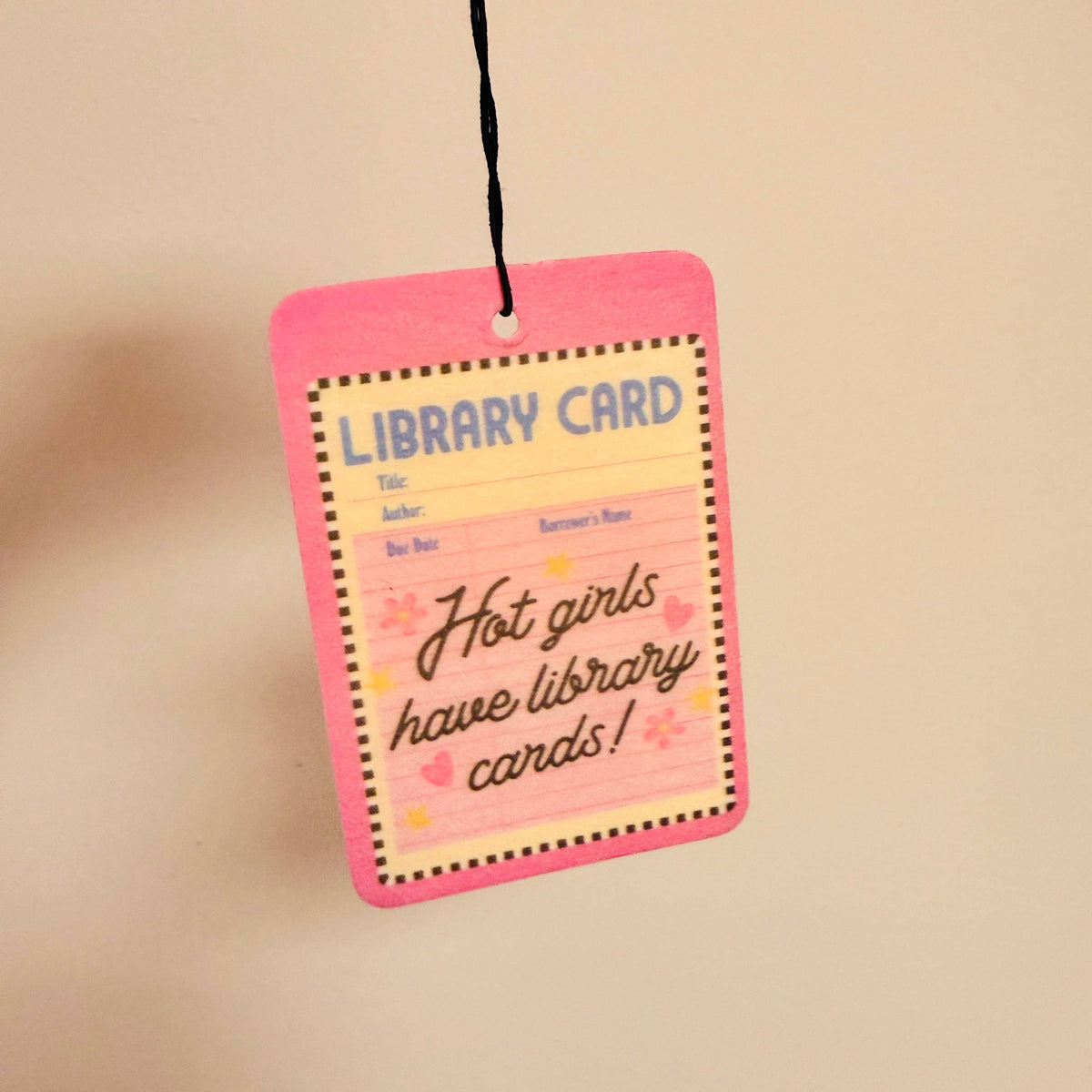 A pink library card tag with a black hanging string, featuring the text 'Hot girls have library cards!' in cursive, set against a plain beige background.