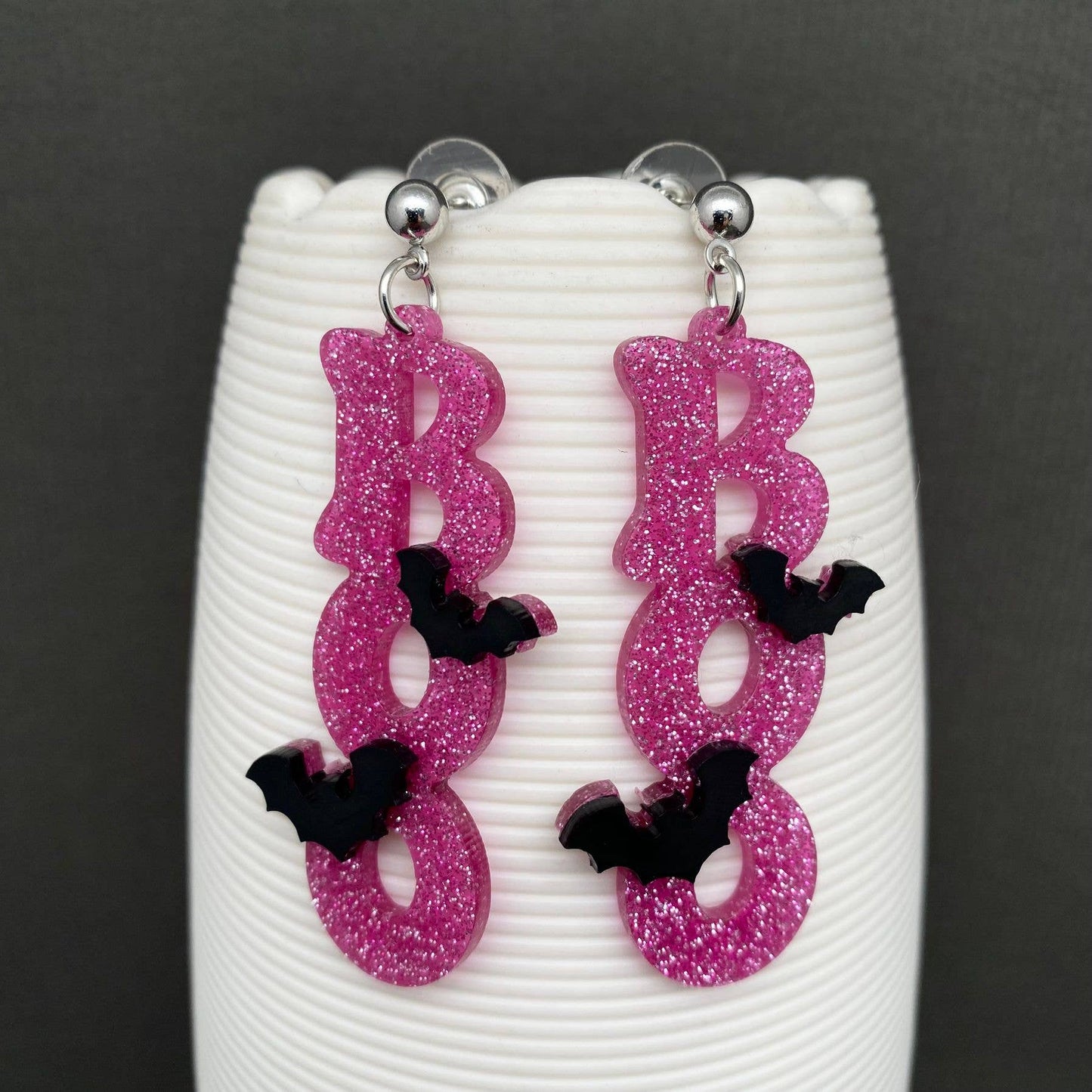 A pair of dangling earrings shaped like the word "BOO" in glittery pink letters, with small black bat accents and silver-tone posts, displayed on a white ribbed stand against a dark gray background.