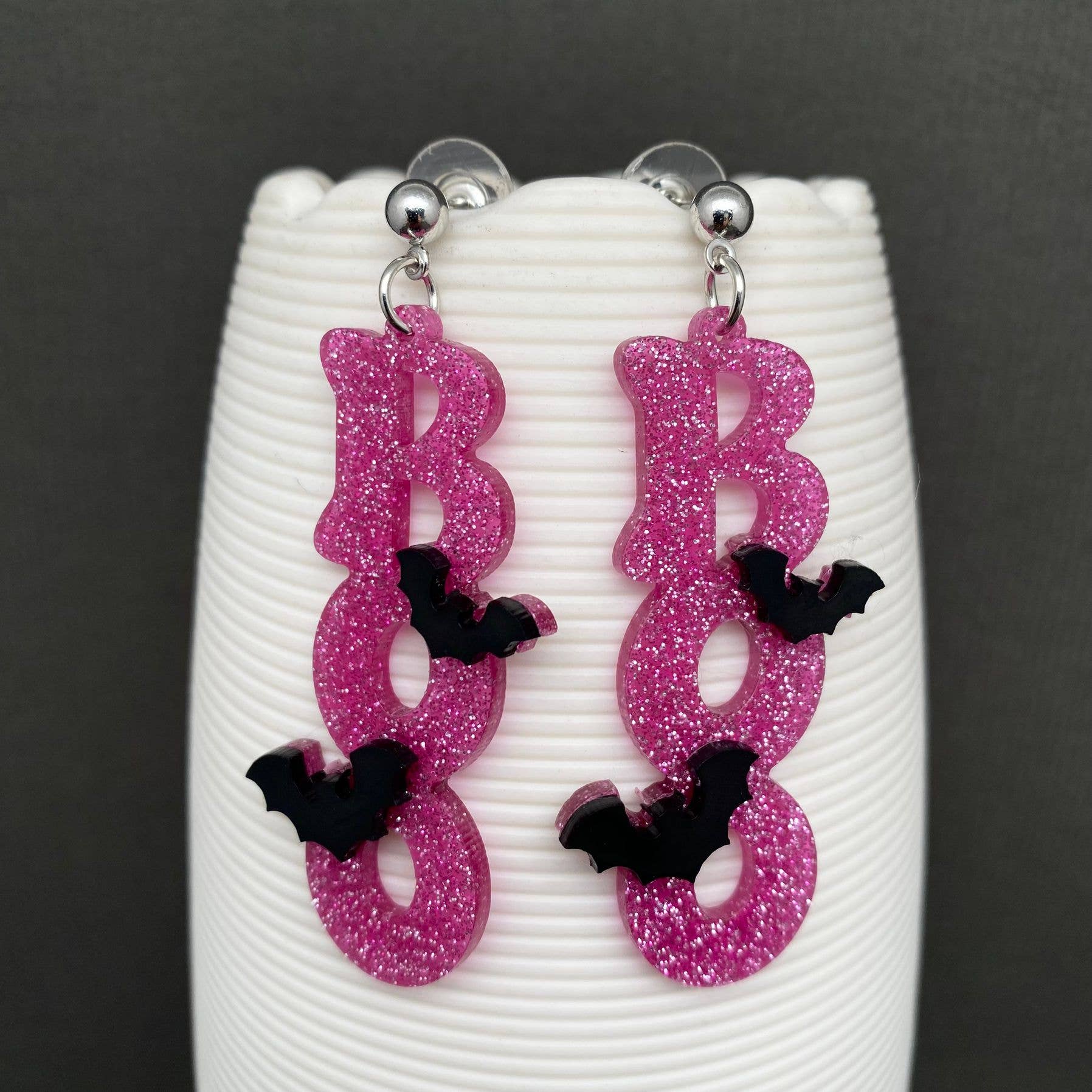 A pair of dangling earrings shaped like the word "BOO" in glittery pink letters, with small black bat accents and silver-tone posts, displayed on a white ribbed stand against a dark gray background.