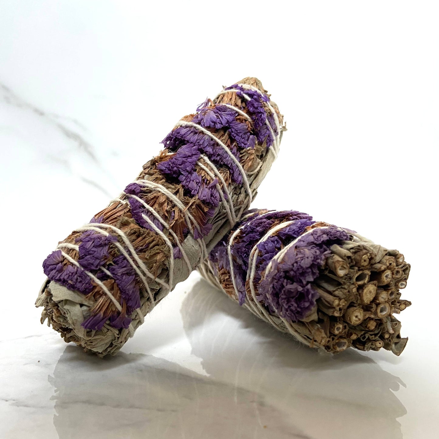 Two bundled sage smudge sticks with purple and white ribbons on a light background