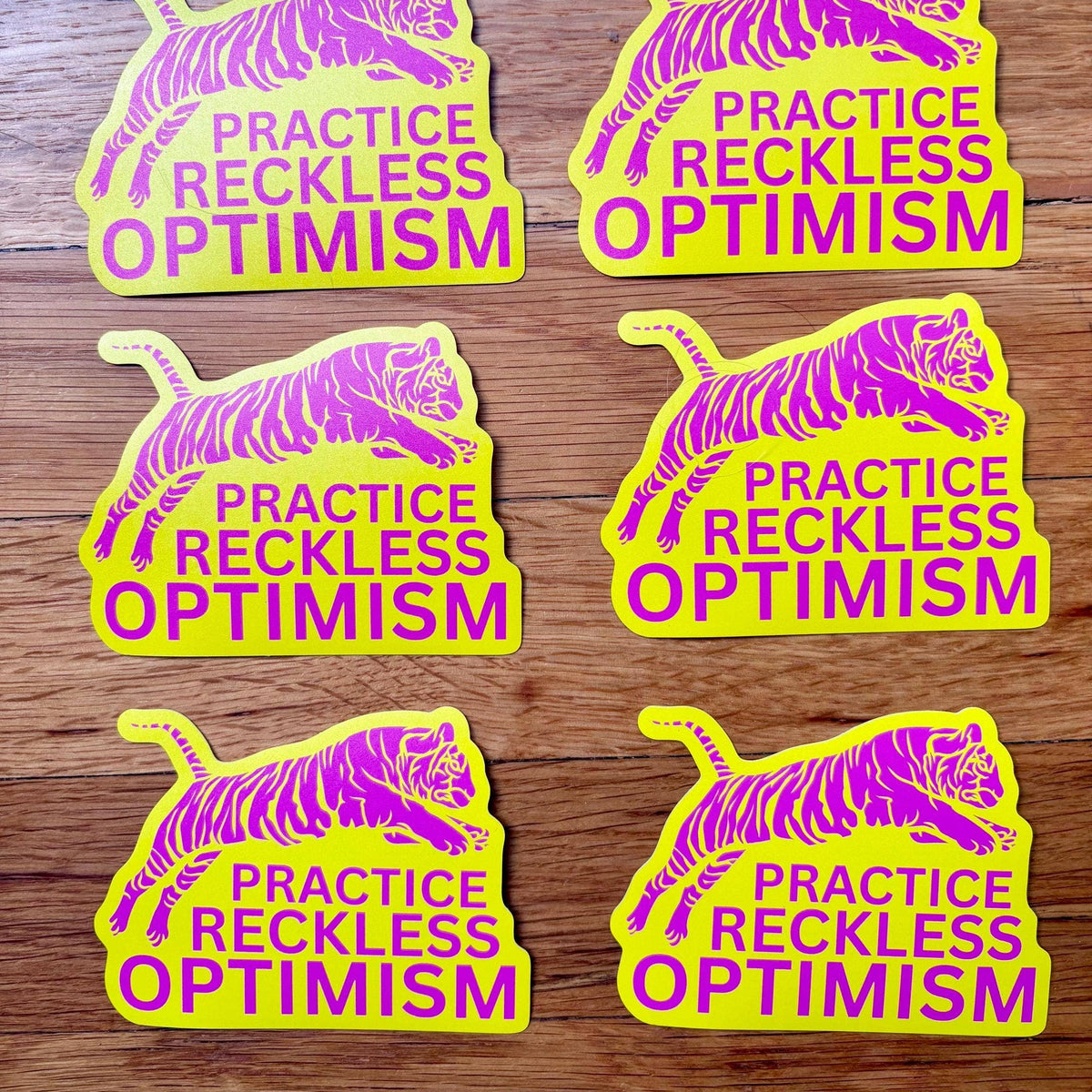 Five stickers with tiger illustrations and 'Practice Reckless Optimism' text on a wooden surface.