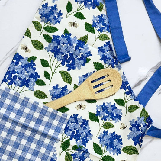 Floral apron with blue hydrangeas and green leaves, wooden spatula, and checkered fabric on a white background.