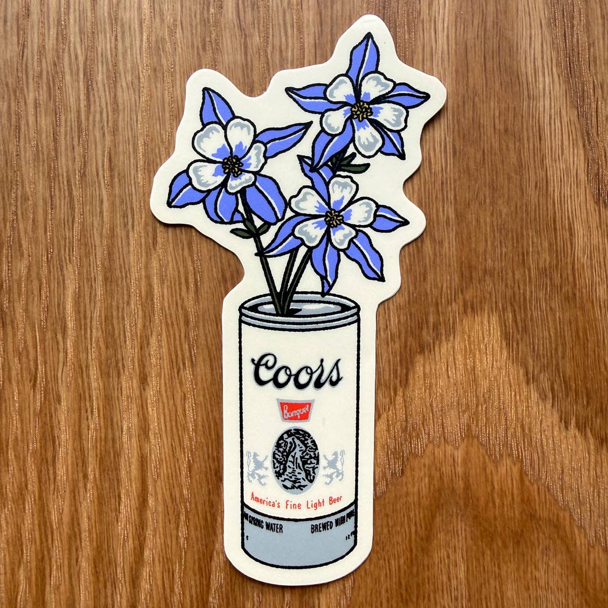 A die-cut sticker featuring blue and white flowers rising from a Coors can, placed on a brown wood surface.