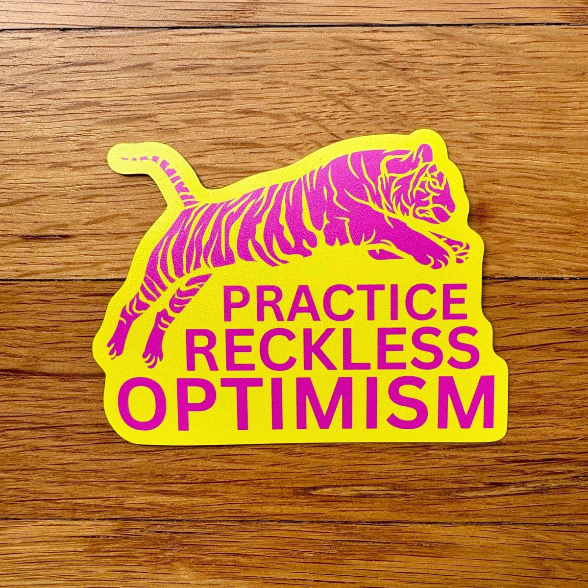 Sticker with a tiger design and text 'Practice Reckless Optimism' on a wooden surface