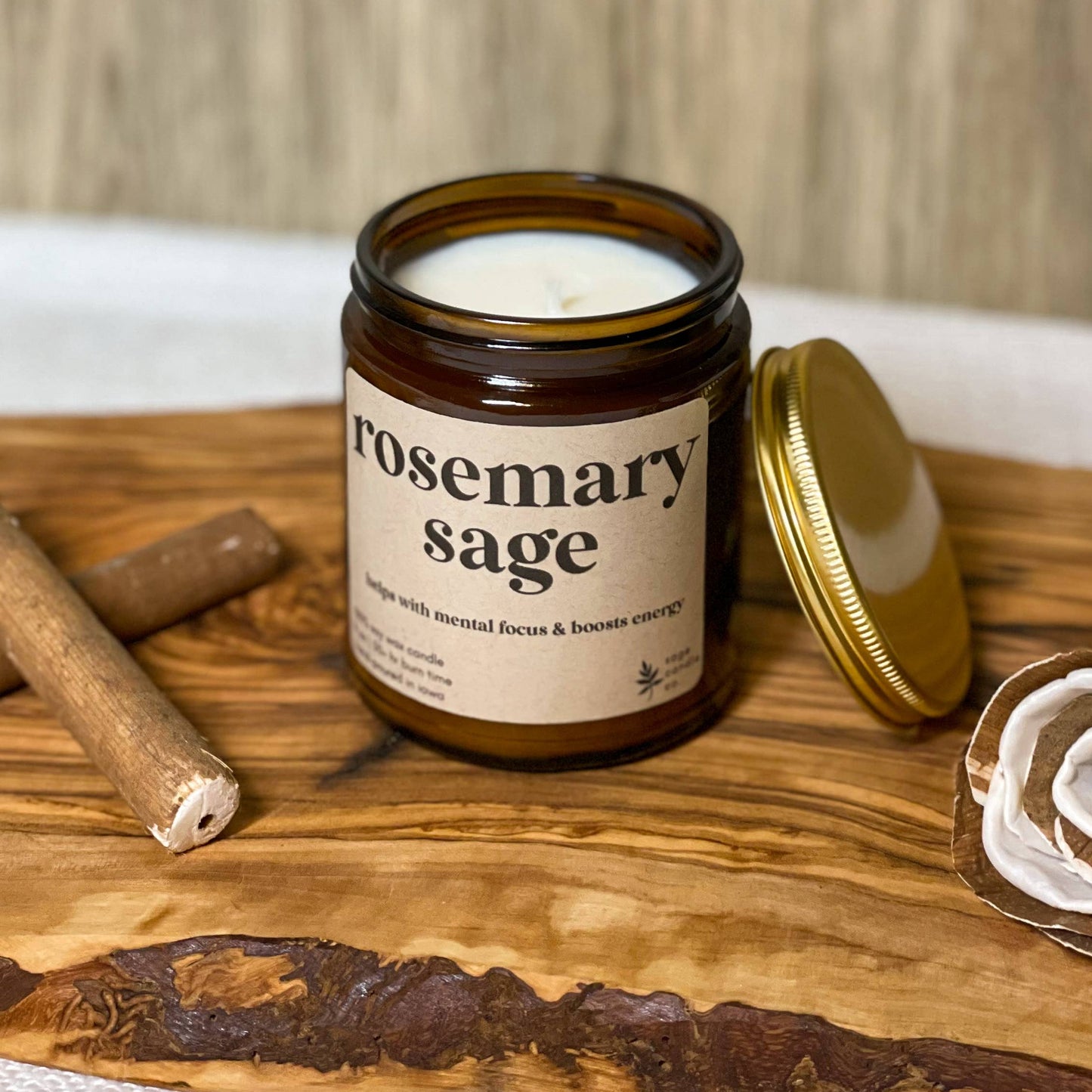 A soy candle in an amber glass jar with a cream label reading 'rosemary sage' and a gold lid resting beside it on a wooden board, styled with cinnamon sticks and a dried rose against a wood plank background.