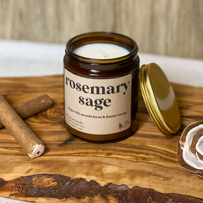 A soy candle in an amber glass jar with a cream label reading 'rosemary sage' and a gold lid resting beside it on a wooden board, styled with cinnamon sticks and a dried rose against a wood plank background.