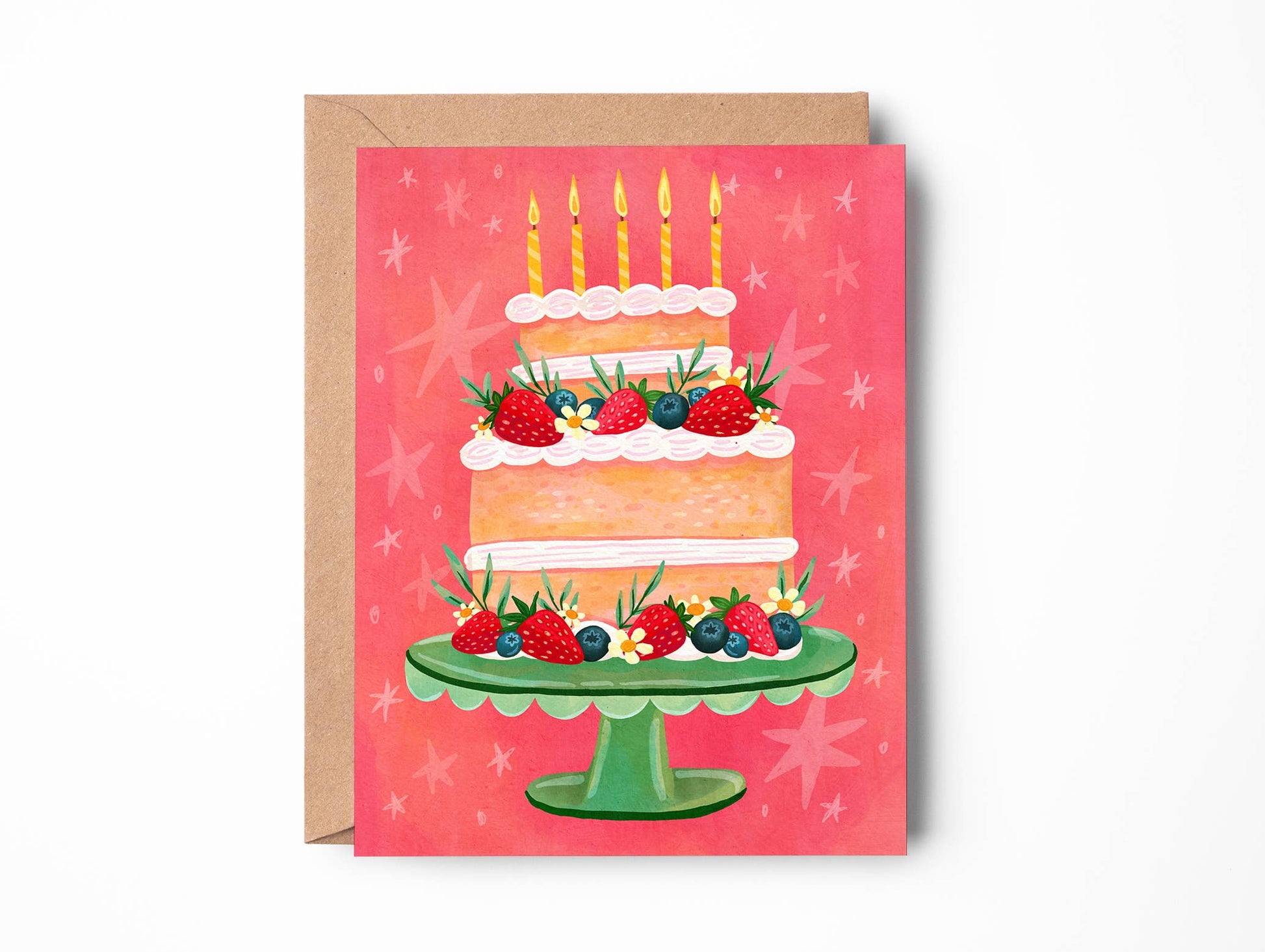 A greeting card featuring an illustration of a three-tiered birthday cake with candles, strawberries, and blueberries on a green stand, set against a pink background with star motifs. The card is shown with a brown kraft envelope behind it on a white background.