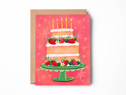 A greeting card featuring an illustration of a three-tiered birthday cake with candles, strawberries, and blueberries on a green stand, set against a pink background with star motifs. The card is shown with a brown kraft envelope behind it on a white background.