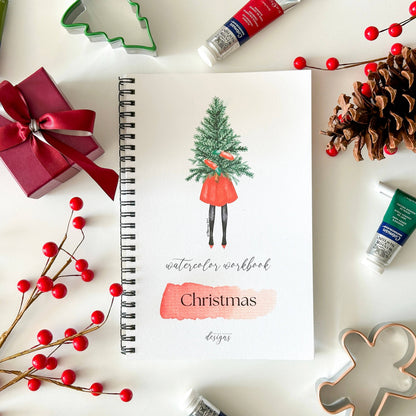 A spiral-bound watercolor workbook titled 'Christmas designs' lies on a white surface, surrounded by holiday props including red berries, a pinecone, a wrapped red gift, and paint tubes. The cover features an illustration of a person carrying a Christmas tree.