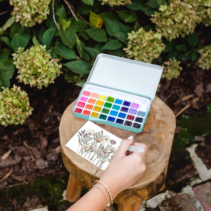 Person painting with watercolors on a small stool outdoors, surrounded by greenery.