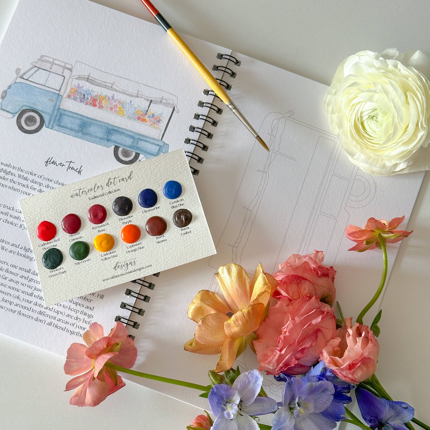 A styled flat-lay of an art journal featuring a watercolor truck illustration, a paintbrush, a color wheel card with multiple paint swatches, and a bouquet of pink, blue, and yellow flowers on a white surface.