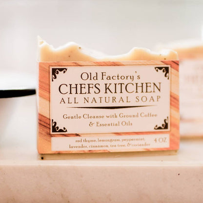 A bar of Old Factory's Chefs Kitchen All Natural Soap sits on a light marble countertop, with its paper packaging box in sharp focus and a second bar blurred in the background.
