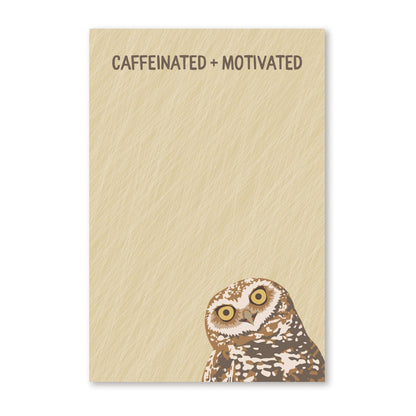 A rectangular notepad with a textured beige cover, printed with the phrase 'CAFFEINATED + MOTIVATED' at the top and featuring a painted owl at the bottom right, shown on a white background.